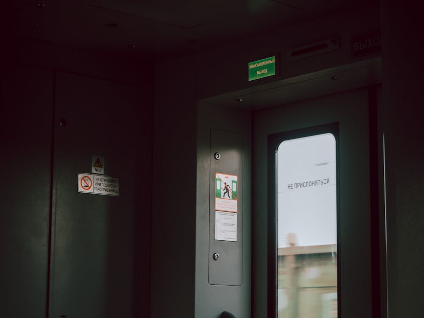A green illuminated emergency exit sign mounted on a dark ceiling, showing a running figure and an arrow pointing to the right. The sign is attached to a metal pipe or conduit and is positioned in an