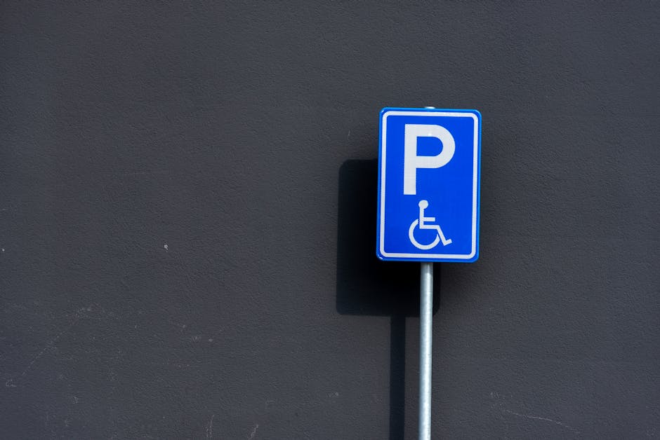 A blue parking sign with a white 'P' and a wheelchair symbol, mounted on a metal pole against a dark grey exterior wall. The sign designates accessible parking in the area, likely adjacent to a proper