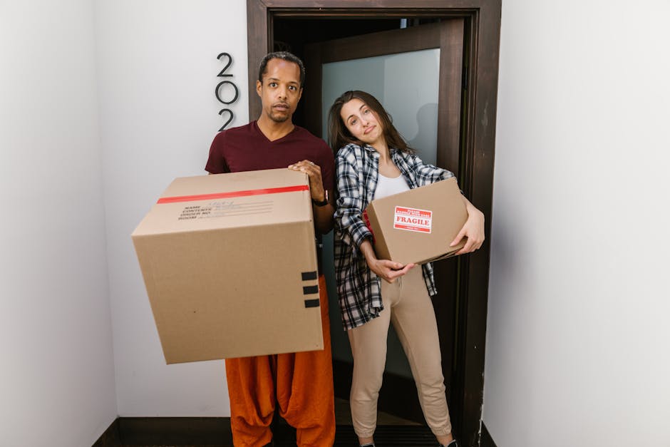 Two individuals, a man and a woman, stand inside a doorway of a residential property, each holding a cardboard box used for home relocation. The man, positioned to the left, appears to have a serious